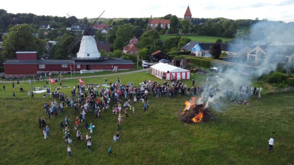 Dronefoto af mange mennesker samlet ved Skt. Hans-bål med den hvide mølle og naturen i baggrunden.